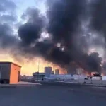 Heavy black smoke engulfing urban sky behind industrial facilities and road network