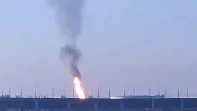 Blazing fire and rising smoke trail visible behind airport perimeter concrete barrier