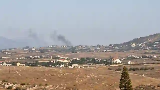 Dark smoke rising into the sky behind rural hillside buildings and open desert terrain