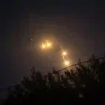 Middle East security summit delays: Glowing illumination flares and smoke trails falling through dark night sky above tree line