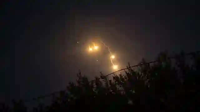 Middle East war crude oil surge: Glowing illumination flares and smoke trails falling through dark night sky above tree line